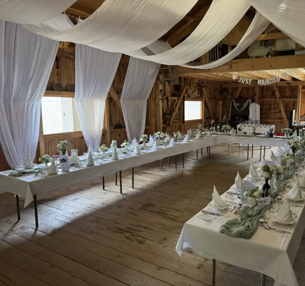 Holzscheune mit weissen Stoffbahnen an der Decke und langen, weiss gedeckten Hochzeitstafeln mit weissen Blumen