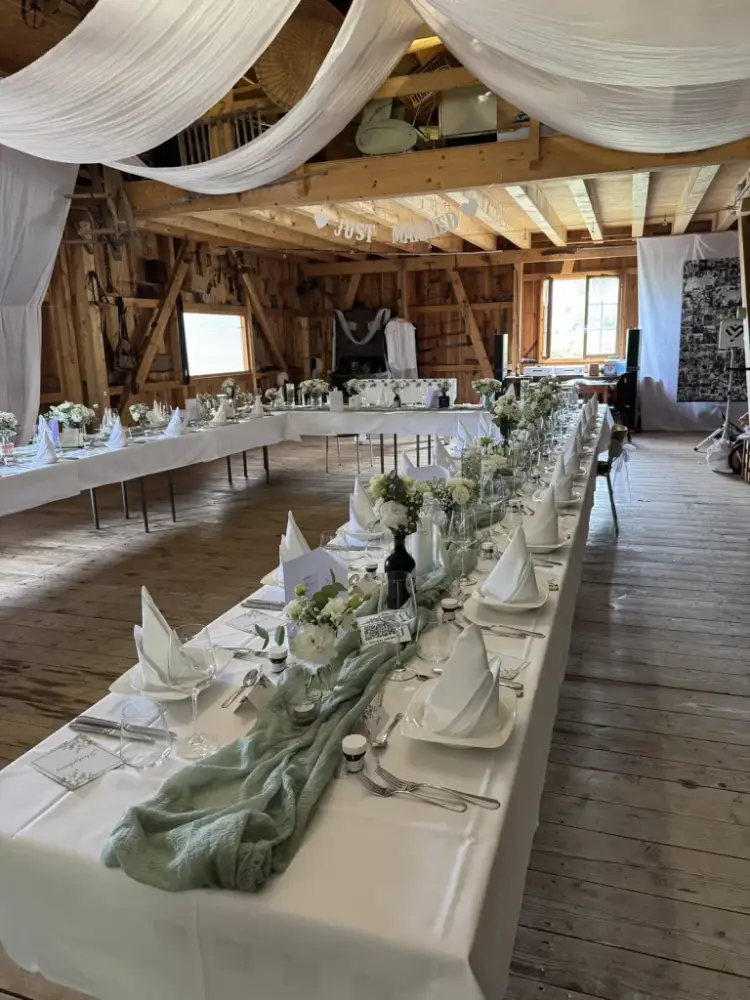 Lange Hochzeitstafel mit salbeigrünem Tischläufer, weissen Blumen und Servietten in der Scheune, darüber Stoffbahnen