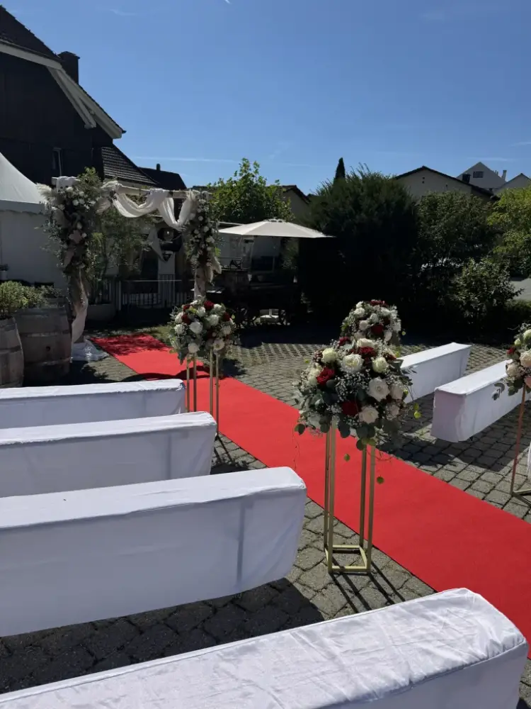 Roter Teppich als Traugang zwischen goldenen Blumensäulen mit roten und weissen Rosen, am Ende der Traubogen mit Tüll
