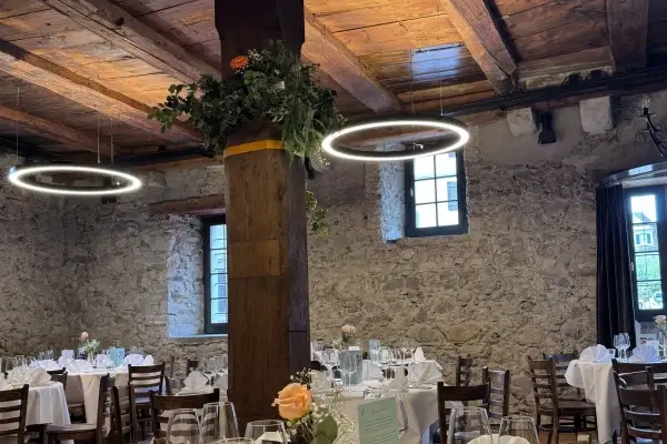 Festlich gedeckte runde Hochzeitstische unter alten Holzbalken und Ringleuchten im Mühleraum des Mühlerama Seon, Holzsäule mit Blumenschmuck und Natursteinwände