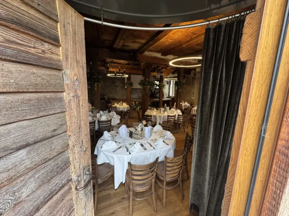 Blick durch das alte Holztor auf runde Hochzeitstische mit weissen Tischtüchern, Holzbalken und Natursteinwänden im Mühleraum