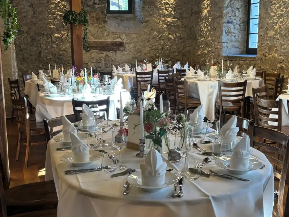 Runde Hochzeitstische mit weissen Tischtüchern, gefalteten Servietten, Stabkerzen und Wildblumen vor der beleuchteten Natursteinmauer im Mühleraum