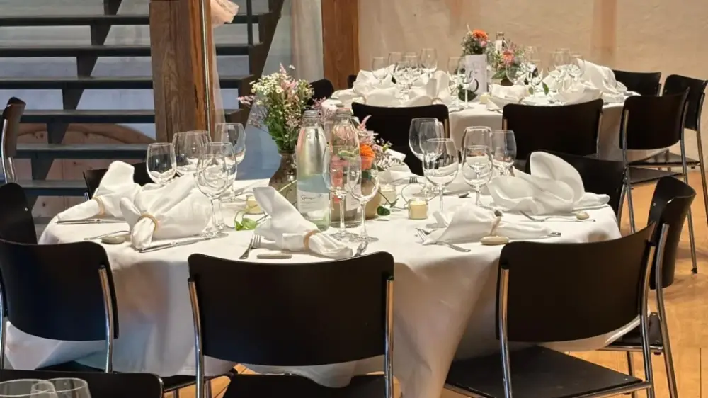 Festlich gedeckte runde Hochzeitstische mit weissen Tischtüchern, Wildblumen und Kerzen im Festsaal mit Holzbalken im Schloss Liebegg