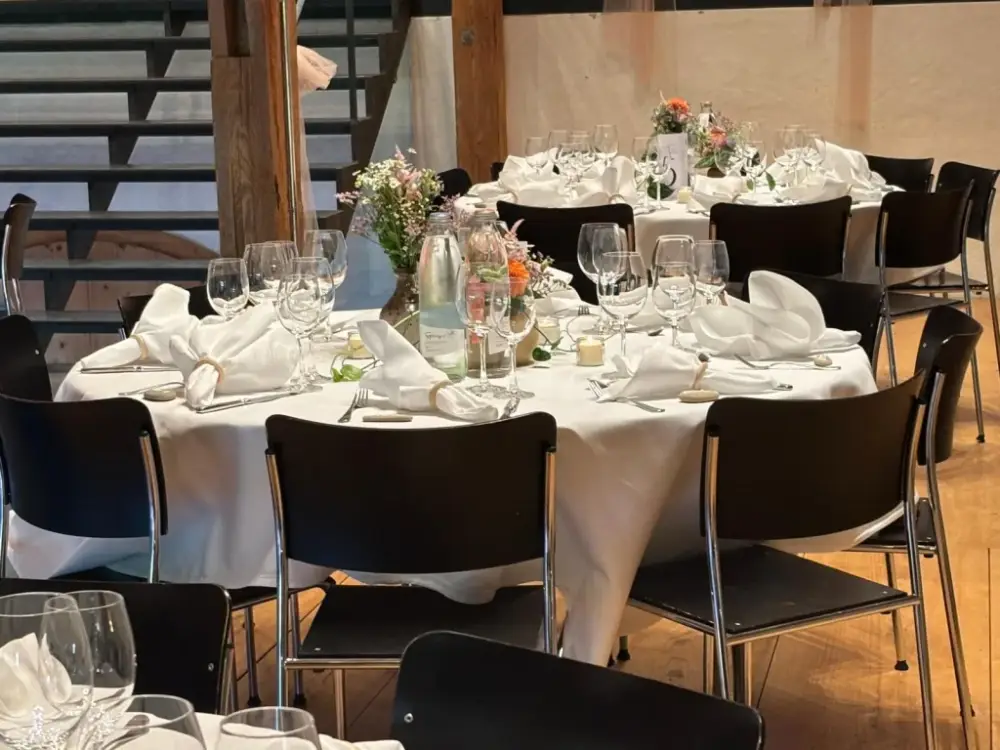 Runde Hochzeitstische mit weissen Servietten, Kristallgläsern und Wildblumen in Keramikvasen vor Holzbalken und Treppe